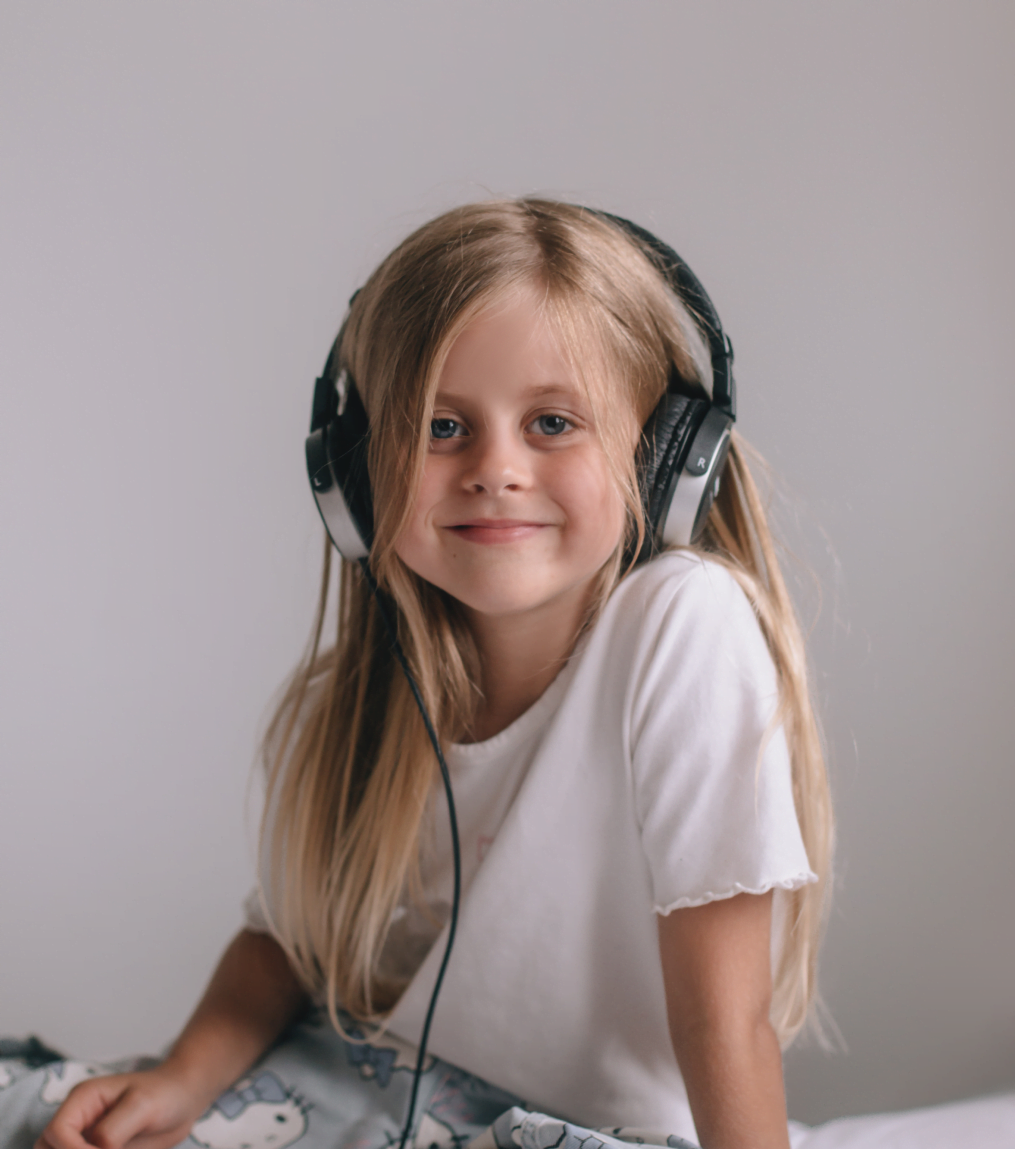 Młoda dziewczyna z długimi blond włosami siedzi w dużych słuchawkach, lekko się uśmiechając. Jest ubrana w białą koszulkę na prostym, jasnym tle.